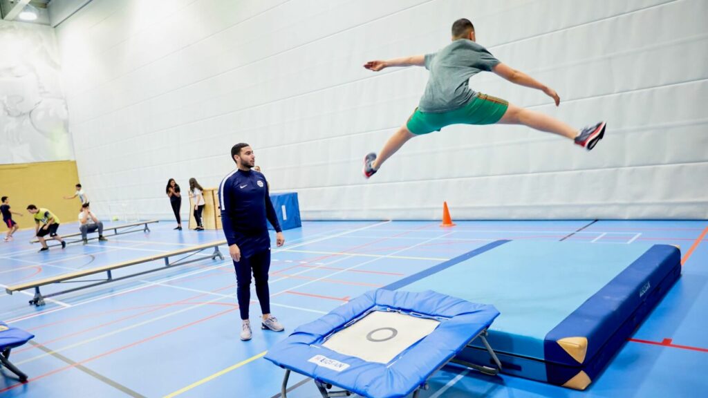 Een leerling springt wijdbeens van de trampoline terwijl de gymdocent toekijkt.
