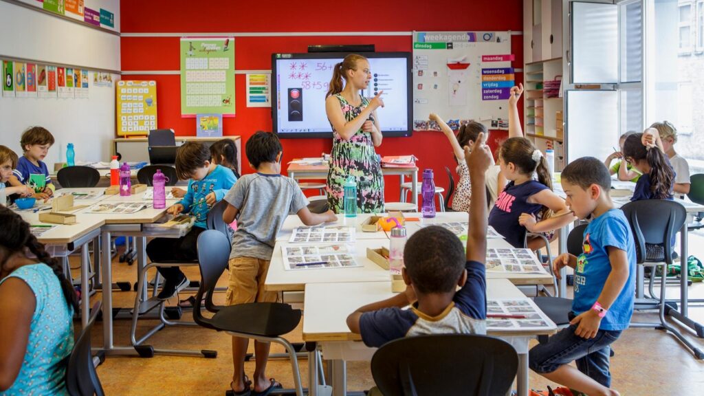 Een juf staat voor de klas met leerlingen die hun vinger opsteken